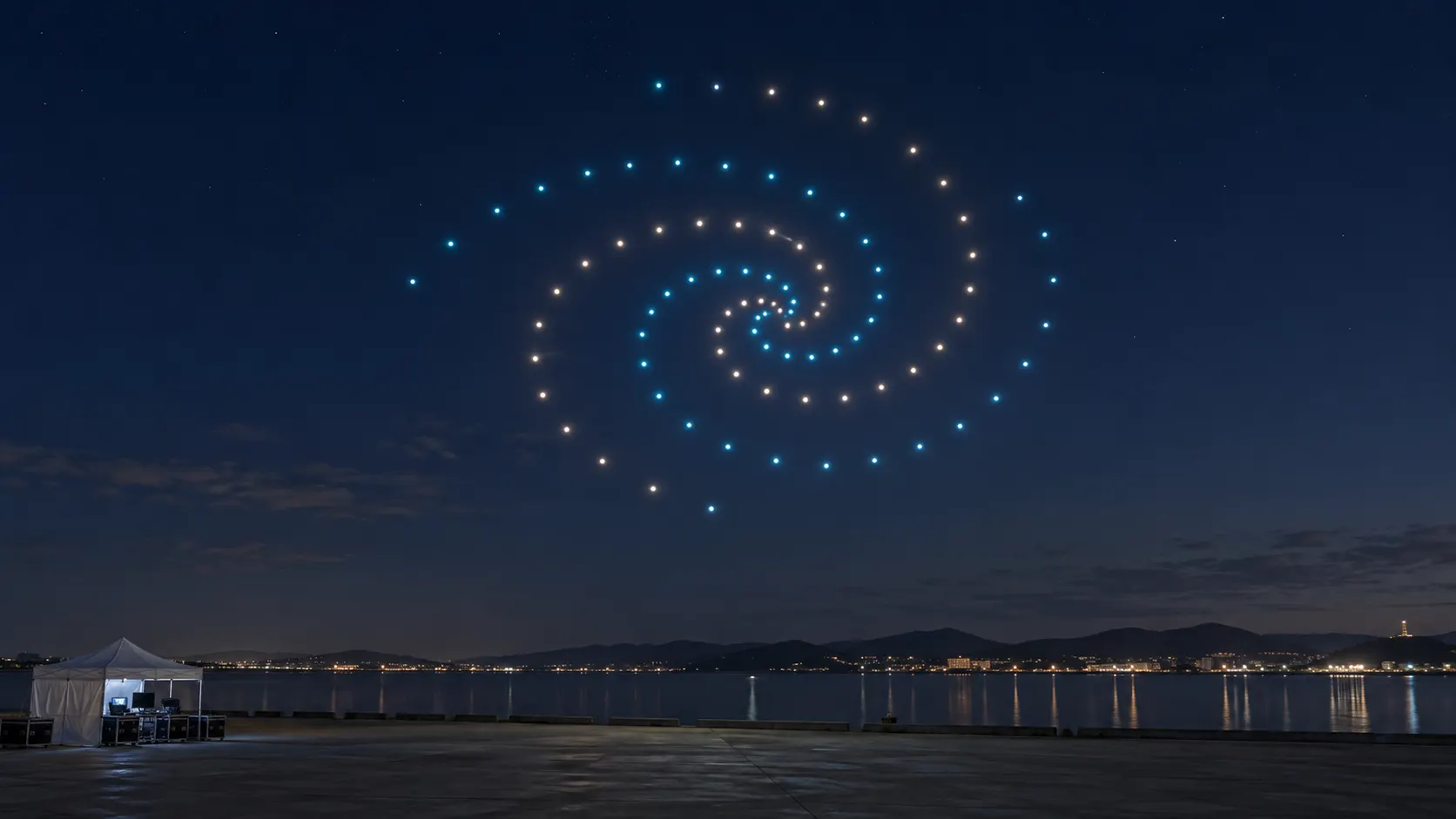 ฝูงโดรนแสดงแสงสีฟ้าและขาวเป็นรูปเกลียวเหนือพื้นที่ริมน้ำในเวลากลางคืน